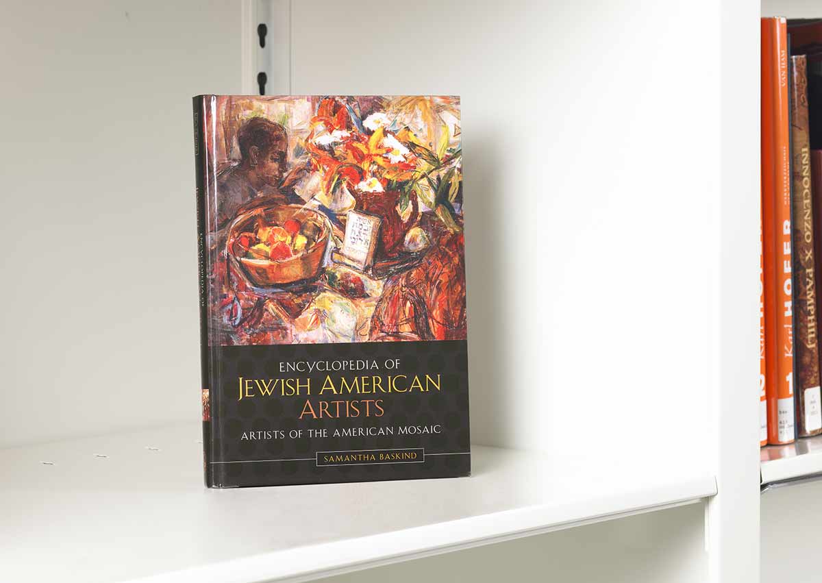 Book on a white shelf whose cover features a figure surrounded by baskets of fruit and a book with Hebrew writing