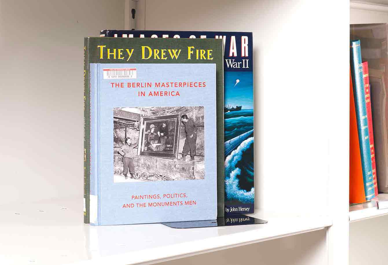Three books stacked on a white shelf. The front book cover features an image of two soldiers recovering an oil painting.
