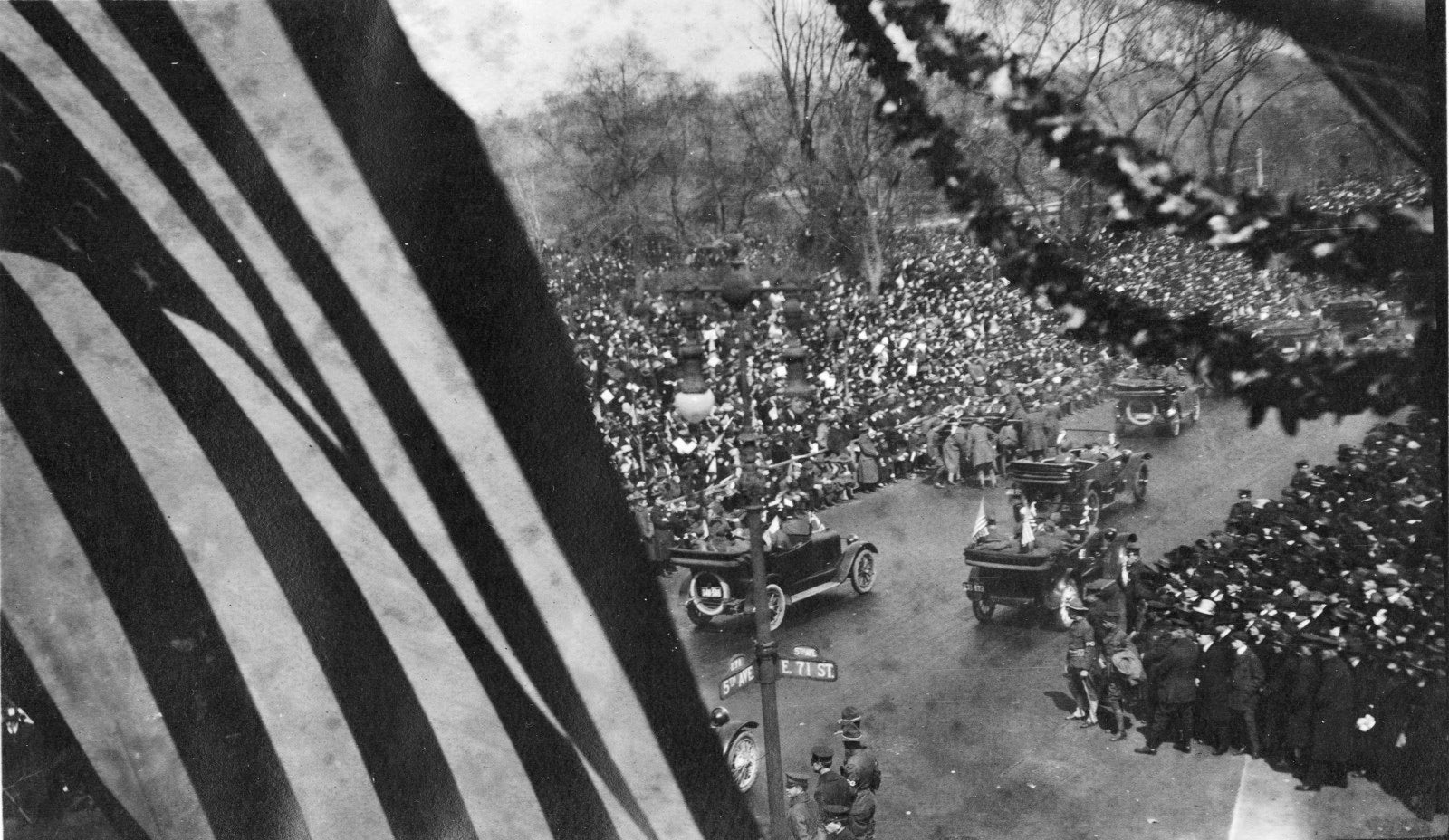 A military parade along Fifth Avenue in New York City, with an American flag and leafy garlands in the foreground