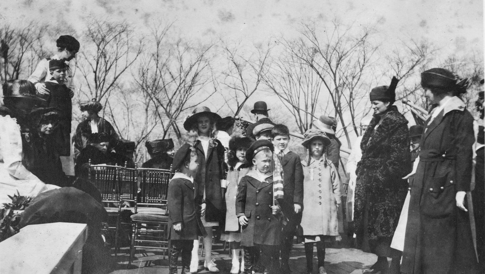 Women and children gathered on a roof set up with benches and chairs. They wear stylish winter clothing and many wear hats. A boy at center holds an American flag.