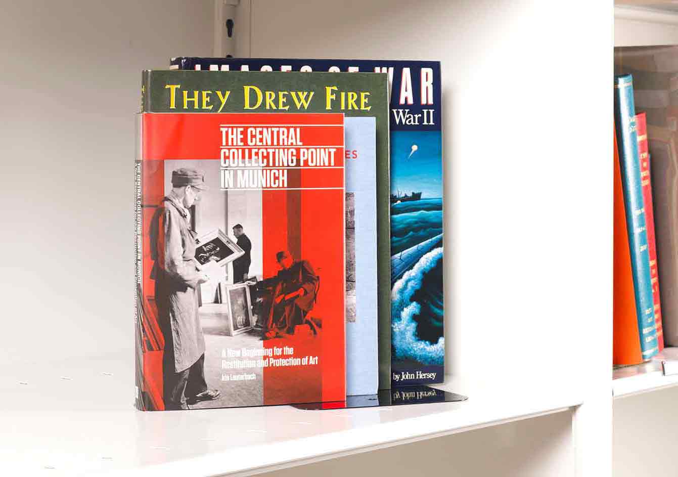 Four books on art and World War Two stacked on a white shelf. The front book cover is orange with an image of army officers handling artworks.