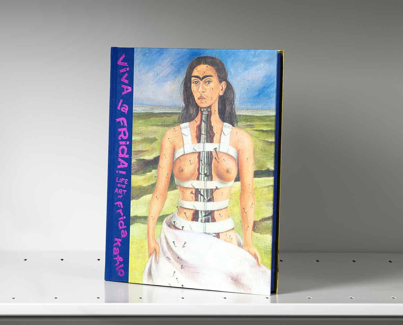 Book on a gray shelf whose cover features a self-portrait of Frida Kahlo wearing white back braces with an interior view of her spine showing