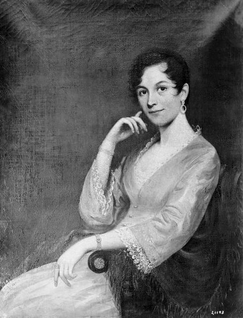 Half-length portrait of a young woman in a dress and long-sleeved jacket seated in a chair.
