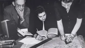 Two women and a man at a desk holding books and marking a map with pencils and rulers