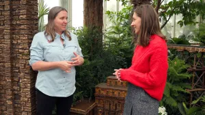 video still of two people talking together in front of model of building