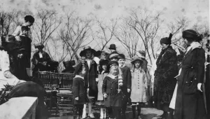 Women and children gathered on a roof set up with benches and chairs. They wear stylish winter clothing and many wear hats. A boy at center holds an American flag.