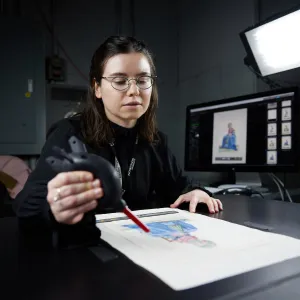 A woman with short dark hair and glasses sits using a tool to digitize a work of art on paper