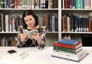 A woman with short dark hair reads a small book at a desk next to a stack of seven other books