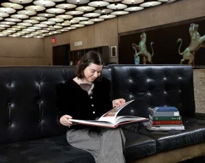 Woman with short brown hair reading a book on a black bench in a museum lobby with a large screen and a ceiling covered in circular lamps