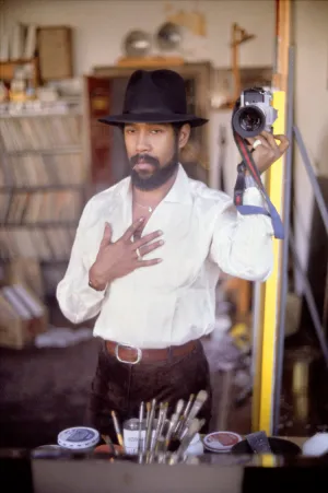 photo of man wearing a black hat holding a camera in one hand, and his other hand on his chest