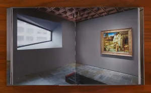 Book on a wooden table open to a spread showing a gray museum gallery with a large trapezoidal window opposite a painting of a saint in nature