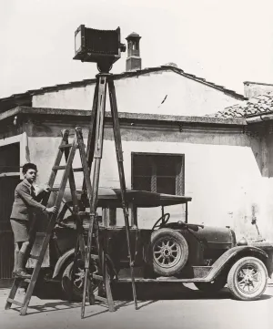 Black and white photograph from 1938. A boy standing on a ladder in front of a car and building. A camera is on a tall tripod in the foreground.