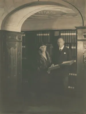 Black and white photograph from circa 1920. A mature man and woman standing in an alcove in front of shelves with archival boxes. The man is holding an open archival box.