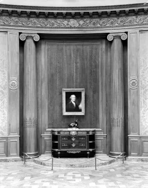 Black-and-white image of a museum gallery displaying a portrait of George Washington, a bronze sculpture, and a chest of drawers