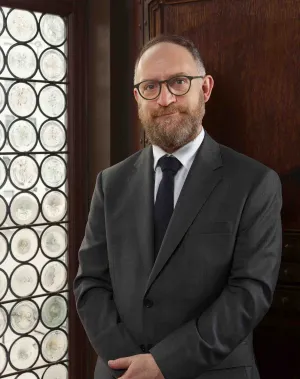 Headshot of Luciano Johnson, new Andrew W. Mellon Chief Librarian, wearing a grey suit with black glasses, dark tie, in a wood paneled room with a rondels stained glass window on the left