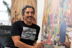 Artist Kent Monkman sitting in front of a colorful painting for his Royal Ontario Museum exhibition