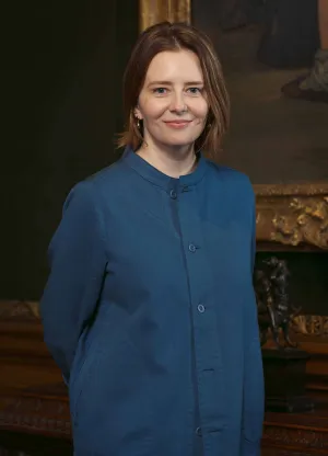 Woman wearing a blue, collarless, button down long sleeve shirt with short brown hair, standing in front of table with part of a painting behind her on the wall