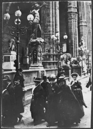 black and white photograph of a group of men and women wearing cloaks and hats walking in front of stone columns and sculptures 