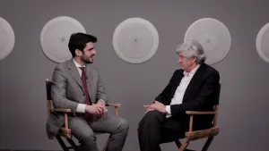 video still of Giulio Dalvit and Giuseppe Penone seated and talking in gallery with disks behind them